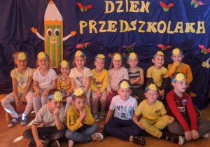 Na zdjęciu widać uśmiechające się dzieci z grupy VI Słoneczka na tle napisu Dzień Przedszkolaka w opaskach na głowach ze słoneczkami